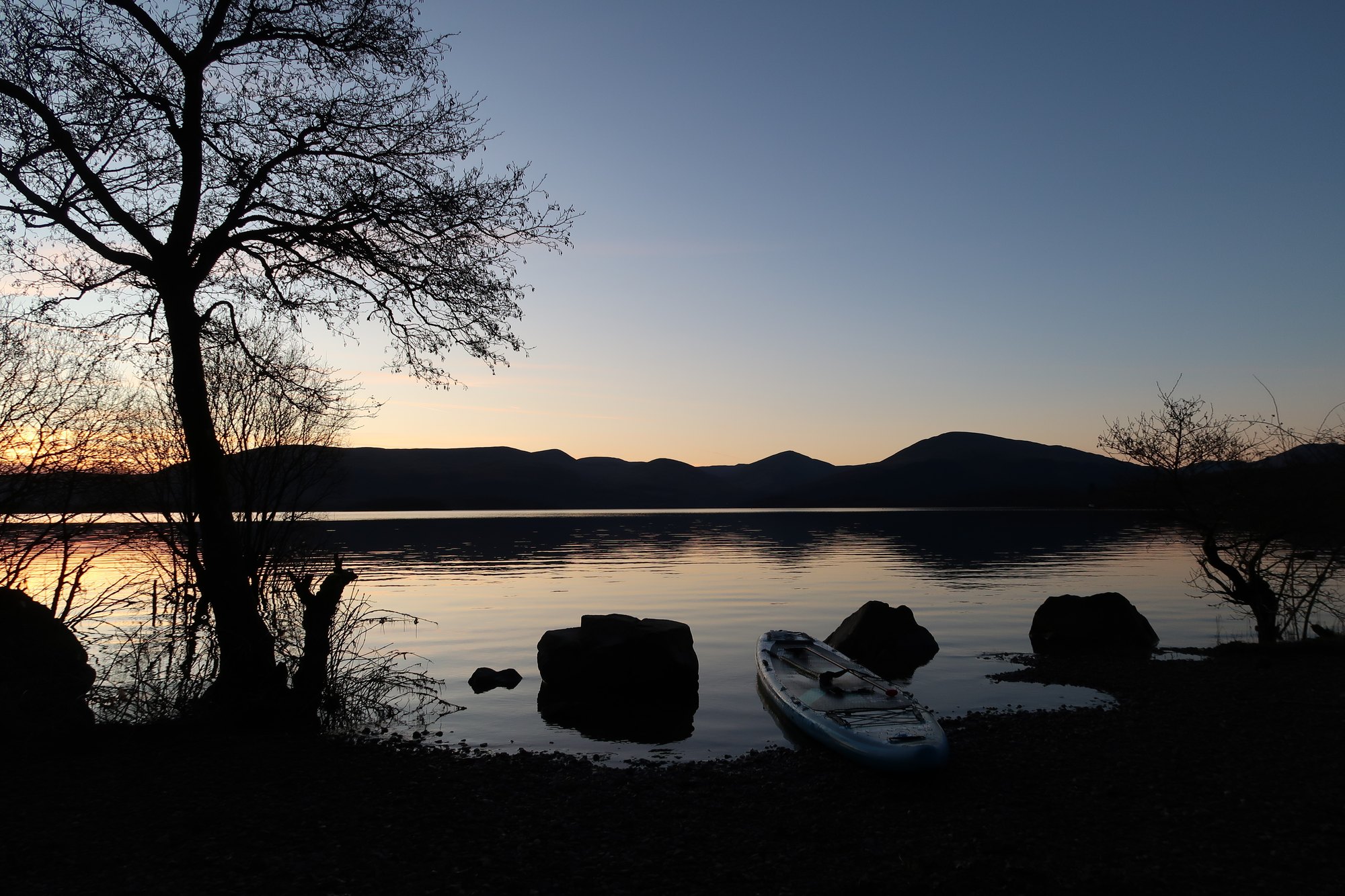 A paddleboard pulled up on the pebble shore of Loch Lomond at dusk, with bare trees against the last light.